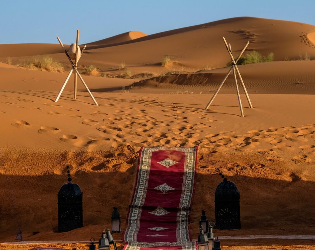 A vertical shot of a red carpet in the middle of lamps and stick tripod with a rock in the distance in the desert
